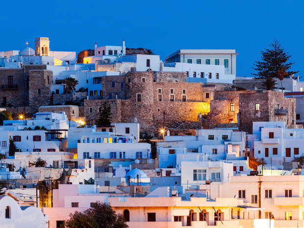 White and stone buildings sit on a hillside, illuminated by twilight. Several properties feature flat roofs and balconies, with some trees interspersed. A church with a small bell tower is visible.